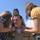 A uniformed military servicemember, wearing a tan/khaki uniform with multiple service ribbons and a badge, is being embraced by two young girls in a joyful reunion scene. A woman stands behind them, appearing to oversee the emotional moment. The setting appears to be an outdoor military installation under a clear blue sky.