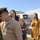 A U.S. Navy enlisted servicemember in khaki uniform adjusts his cover (hat) while smiling, surrounded by what appears to be his family — a woman and two young girls — in an outdoor military base setting. The scene appears to be a celebratory or ceremonial occasion, possibly a promotion, homecoming, or award ceremony. The location appears to be a NATO or U.S. military installation in Italy, based on the bilingual signage visible on a nearby vehicle.