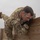 Ivy Week 2022 Mabry Mile: Up and Over A male soldier in U.S. Army OCP (Operational Camouflage Pattern) uniform is scaling or climbing over a wooden wall obstacle. He is wearing black tactical gloves and has a U.S. flag patch and what appears to be a unit patch on his left sleeve. The image appears to be taken during a physical fitness or obstacle course training event under overcast skies.