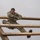 Ivy Week 2022 Mabry Mile: Keep Climbing! A soldier in camouflage uniform is navigating a wooden obstacle course structure, appearing to climb or traverse over a ladder-like wooden frame. The individual is exerting significant physical effort, with arms braced over the top rail and legs spread across lower rungs. Mud and debris are visible on the structure and the soldier's clothing, suggesting an outdoor military training exercise.