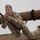 Ivy Week 2022 Mabry Mile: Sliding on a rope A military soldier in full camouflage uniform is navigating a rope obstacle course, gripping a black rope while suspended horizontally between wooden log beams. The soldier displays intense physical exertion and concentration, with mud-covered boots visible. This appears to be a military physical training or obstacle course exercise.