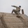 Ivy Week 2022 Mabry Mile: Slide down swiftly Two individuals in military camouflage uniforms and black knit caps are navigating a large wooden obstacle wall, consistent with a military training course. One person is cresting the top of the inclined wooden structure while another assists or follows from behind. The overcast sky and muddy conditions on the wall suggest cold, wet weather during the exercise.