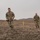 Ivy Week 2022 Mabry Mile: Keep running! Two U.S. Army soldiers in MultiCam/OCP camouflage uniforms are running or jogging across a barren, arid terrain. The foreground soldier has a visible name tape and U.S. Army tape on his uniform, and appears to be holding gloves in one hand. The background is characterized by flat, dry ground with low hills and an overcast sky.