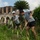 A group of people in matching gray t-shirts and black shorts are performing grounds maintenance work, clearing overgrown vegetation near ancient Roman ruins. The ruins in the background feature characteristic Roman arched architecture with brick construction, suggesting this may be an archaeological site in Italy. The workers appear to be engaged in a volunteer or organized cleanup effort at a historical monument.
