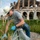 A man wearing a white cowboy hat, blue sunglasses, gray t-shirt, and jeans is performing cleanup or weeding work in an overgrown area near ancient Roman ruins. He is wearing blue latex gloves and collecting vegetation into a clear plastic bag. The background features a large, well-preserved ancient Roman amphitheater or arena structure with brick arches and stonework.
