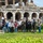 A group of approximately 18-20 people pose for a group photo in front of what appears to be the Anfiteatro Campano (Capuan Amphitheater) in Santa Maria Capua Vetere, Italy, an ancient Roman ruin. The group is holding rakes and gardening tools, suggesting they participated in a volunteer cleanup or landscaping activity at the historic site. The setting includes well-maintained green grass and hedges in the foreground with the ancient stone arched structure prominently visible behind them.