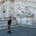 A male tourist is photographing a large classical reclining marble statue on the exterior of a grand white stone building, likely the Palazzo Senatorio on Capitoline Hill in Rome. The statue depicts a reclining river god figure, characteristic of ancient Roman sculptural tradition. An Italian flag is visible in the background, confirming the Roman civic setting.