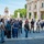 A tour group of approximately 20-30 people is gathered on the Piazza del Campidoglio in Rome, Italy, listening to a guide holding books or materials on the right side of the frame. The iconic equestrian statue of Marcus Aurelius is visible in the background, along with the facade of the Musei Capitolini (Capitoline Museums), identified by a red banner. The scene appears to be a daytime guided educational or tourist tour under clear skies.