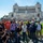 A group of approximately 18-20 tourists poses for a group photo in front of the Altare della Patria (Vittoriano monument) in Rome, Italy. The neoclassical white marble monument is clearly visible in the background, along with an Italian flag flying from its colonnade. The group appears to be on an organized tour, as evidenced by visible lanyards and name badges worn by several members.