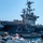 A group of people aboard what appears to be a tour or ferry boat are viewing a large US Navy aircraft carrier at close range from the water. The carrier's island superstructure is prominently visible with radar arrays, antennas, and the hull number '75' partially visible. Multiple aircraft can be seen parked on the carrier's flight deck.