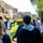 A tour guide wearing a blue jacket and orange lanyard badge is addressing a group of tourists at what appears to be an ancient archaeological site, likely Pompeii based on the visible ruins and architectural remains. The guide is gesturing with one raised hand while speaking to approximately 8-10 visitors gathered around him. The setting features well-preserved ancient stone walls, green vegetation, and scaffolding visible in the background indicating ongoing restoration work.