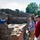 A tour guide in a blue jacket gestures while explaining an ancient Roman thermopolium (food counter) to a group of tourists at what appears to be the archaeological site of Pompeii, Italy. The stone counter features characteristic circular dolia (storage vessels) embedded in the surface, typical of Roman fast-food establishments. The group of approximately 8-10 visitors, some wearing lanyards, listens attentively against a backdrop of ancient ruined walls and trees.