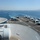 An aerial view from the island superstructure of a US Navy aircraft carrier underway at sea, showing the flight deck populated with multiple fixed-wing aircraft and rotary-wing aircraft. The carrier appears to be departing or operating near a coastal city visible on the horizon. The flight deck shows active deck operations with personnel and ground support equipment visible.