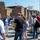 A tour group of approximately 15-20 people is gathered at an ancient Roman archaeological site, likely Pompeii, listening to a guide in a blue jacket who is gesturing and speaking to the group. The ruins consist of exposed brick walls, stone-paved streets, and partially standing structures typical of Roman architecture. A construction crane is visible in the background near a white building, indicating ongoing restoration or excavation work.
