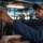 A sailor in navy blue coveralls and a tan baseball cap operates communications or navigation equipment on what appears to be the bridge of a U.S. Navy vessel. Multiple crew members are visible in the background, suggesting an active watch or operational period. The setting is consistent with a naval ship's pilothouse or combat information center.