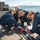 Four military personnel in dark blue coveralls and tan caps are crouched on what appears to be a ship's deck, working on a track or rail mechanism. They are wearing blue latex gloves and handling red cloth material and plastic bags near a recessed deck rail. A large body of water and a prominent volcanic mountain are visible in the background, consistent with the appearance of Mount Fuji near Yokosuka, Japan.
