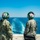 Two U.S. Navy personnel in camouflage uniforms stand at the stern of a naval vessel, looking out over the ship's wake on open blue water. Both wear green flight deck helmets with communications headsets, with visible markings '3T', 'HOLCOMB' on the left individual, and 'SP' on the right. A coastline with urban development is faintly visible on the left horizon.