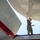 A military service member in U.S. Navy working uniform (NWU Type III camouflage) is performing maintenance on a large aircraft, applying a substance to the underside of what appears to be a radome or fuselage section. The photo is taken from a low angle looking upward, giving a dramatic perspective of the worker reaching overhead. The number '600' is visible on the aircraft surface, and red and white paint markings are also present.