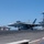 A U.S. Navy F/A-18 Super Hornet (bureau number 407) is being catapult-launched from the flight deck of a Nimitz-class aircraft carrier. Steam from the catapult system is visible beneath the aircraft as it prepares for takeoff. Multiple other aircraft and flight deck personnel in colored jerseys are visible in the background.