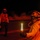 A night operations scene on what appears to be a naval aircraft carrier flight deck. A crew member in the foreground wearing a helmet with ear protection, goggles, and a jacket labeled 'FLY' is holding two illuminated light wands used for aircraft marshaling. At least two additional personnel are visible in the background under red ambient lighting.