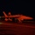A U.S. Navy jet aircraft, marked with the number '501', is positioned on what appears to be an aircraft carrier flight deck at night. The scene is illuminated by intense red/orange lighting, casting a dramatic glow over the aircraft and deck surface. The aircraft appears to be armed with munitions and is likely staged for a night launch operation.