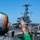 A U.S. Navy aircraft carrier flight deck scene showing flight operations in progress. A crew member wearing a green jersey and orange-striped helmet is visible in the foreground, signaling toward a jet aircraft numbered '502' positioned near the carrier's island superstructure. Multiple deck crew members in various colored jerseys are visible performing their assigned roles.