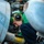 A military aviation mechanic or technician is performing maintenance work inside an aircraft, likely on a naval vessel given the green jersey and helmet indicating a specific deck crew role. The technician is closely inspecting or working on mechanical components within a tight access area of the aircraft. The image is taken from a close, narrow angle between aircraft components, giving a confined, immersive perspective.