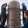 Four naval personnel in dark blue coveralls are working on a large cylindrical weapons system aboard a naval vessel at sea. The equipment appears to be a Phalanx Close-In Weapon System (CIWS) or similar naval defense system, identifiable by its characteristic domed top and riveted cylindrical housing. The sailors appear to be conducting maintenance or inspection procedures on the open ocean.