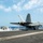 A U.S. Navy fighter jet, marked '201', is captured mid-launch from an aircraft carrier flight deck, with steam from the catapult system visible behind it. The aircraft appears to be an F/A-18 Super Hornet in the process of a catapult-assisted takeoff. A second aircraft and flight deck personnel are visible in the background.