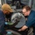 Two naval personnel are performing maintenance on a deck-mounted weapon system aboard a warship at sea. The component labeled 'ENTRANCE END' is visible on what appears to be a close-in weapon system or autocannon feed mechanism. One sailor wears blue nitrile gloves and a tan cap, while the other wears glasses and a dark blue uniform.