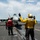 Flight deck personnel are conducting aircraft handling operations aboard a naval aircraft carrier. A yellow-shirted aircraft director is signaling with a raised hand toward an F/A-18 Hornet or Super Hornet positioned on the catapult track. Multiple crew members in color-coded jerseys are visible performing their respective roles on the flight deck.