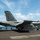 A U.S. Navy F/A-18 Super Hornet (modex 201) is being launched from an aircraft carrier flight deck, with a shooter/catapult officer in a red jersey giving the launch signal. The aircraft bears the 'AB' tail code and checkerboard markings consistent with CVW-7, and carries external stores including what appears to be a centerline fuel tank. A second aircraft (modex 301) is visible in the background on the left side of the deck.