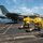 A U.S. Navy carrier-based jet aircraft, identifiable as an F/A-18 Hornet or Super Hornet, is being launched from an aircraft carrier flight deck via catapult. Two crew members wearing yellow 'SHOOTER' jerseys and helmets are crouched in the characteristic launch signal posture. A third crew member in a green jersey is visible at the right edge of the frame.