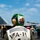 A U.S. Navy carrier deck scene showing personnel from squadron VFA-11 (partially visible on the vest) on what appears to be an aircraft carrier flight deck. The foreground figure wears a distinctive green helmet with a cartoon mascot and the callsign 'Ripper' painted on it. An F/A-18 Super Hornet jet aircraft bearing the number '100' is visible in the background, surrounded by flight deck crew in various colored jerseys.