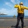 A U.S. Navy flight deck director, identifiable by their yellow jersey and helmet, is signaling on an aircraft carrier flight deck. The director, whose vest reads 'CASH', is directing an F/A-18 Hornet (bureau number 503) on the carrier deck. Additional flight deck personnel are visible in the background near the ocean horizon.
