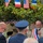 An outdoor ceremony featuring French and American flags displayed together on bunting, suggesting a Franco-American commemorative or diplomatic event. A man wearing a French tricolor mayoral sash (écharpe tricolore) is speaking into a microphone, indicating an official municipal address. The presence of military uniform, musical instruments, and flag decorations suggests a formal public ceremony, possibly related to WWII commemoration or a town twinning event.