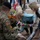 A memorial ceremony is taking place where children are laying flowers at a tribute site surrounded by floral arrangements. A U.S. Army soldier in camouflage uniform with an American flag patch is seated nearby observing. Both a Union Jack (British) flag and an American flag are visible in the background, suggesting an Allied commemorative event, likely related to WWII remembrance.