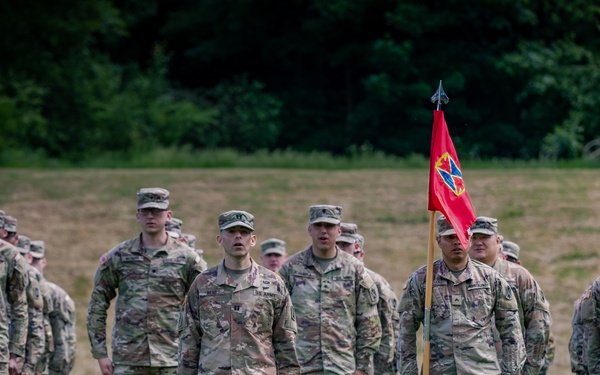 A formation of U.S. Army soldiers in OCP (Operational Camouflage Pattern) uniforms and patrol caps are standing at attention on a grass field. A red unit guidon flag with a distinctive insignia is visible being carried by one of the soldiers. The scene appears to be a formal military ceremony or formation event.