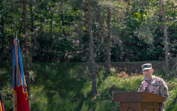 A U.S. Army soldier in camouflage uniform speaks at a wooden podium during an outdoor military ceremony. Three flags are displayed to the left — the U.S. flag, the German flag, and what appears to be a U.S. military unit guidon — mounted on weighted bases on a well-maintained grass lawn. The setting appears to be a formal military event, likely at a U.S. installation in Germany.