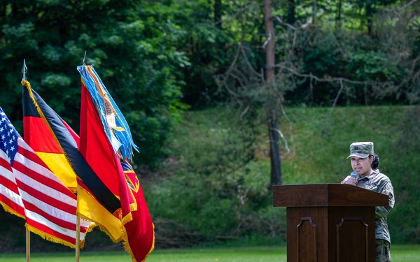 A military service member in camouflage uniform stands at a wooden podium speaking at an outdoor ceremony on a well-maintained grass lawn. Multiple flags are displayed on wooden bases to the left, including two U.S. flags, a German national flag, and at least two military unit/organizational flags. The setting appears to be a formal bilateral or joint military ceremony, likely held at a U.S. military installation in Germany.