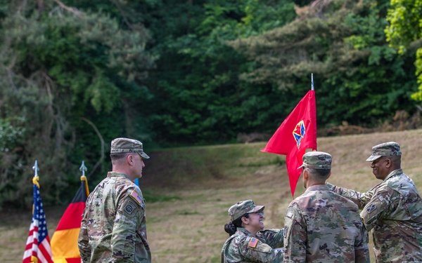 A U.S. Army military ceremony is taking place outdoors on a grass field, likely a change of command or activation ceremony. Several soldiers in MultiCam/OCP camouflage uniforms are participating in a guidon (unit flag) transfer, with both the American flag and German flag visible in the background, indicating this ceremony is taking place in Germany. The red guidon being passed features a distinctive unit insignia consistent with a U.S. Army headquarters element.