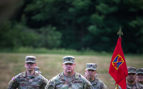 A group of U.S. Army soldiers in Operational Camouflage Pattern (OCP) uniforms are standing in formation on an outdoor field. A central figure identified by nametape as 'O'CONNOR' stands at the forefront, with a soldier to his right holding a red unit guidon featuring a yellow insignia. The scene appears to be a formal military ceremony or formation event.