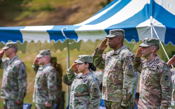 A group of U.S. Army soldiers in OCP (Operational Camouflage Pattern) uniforms are rendering a salute during a formal military ceremony held outdoors. The event is taking place on a grass field beneath a blue and white striped ceremonial tent. Multiple soldiers of varying ranks and genders are visible, standing in formation and saluting in unison.