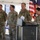 Four military personnel in OCP (Operational Camouflage Pattern) uniforms are gathered under a white tent at what appears to be an official ceremony or groundbreaking event. A female soldier is speaking at a podium/lectern while three male soldiers stand at attention beside her. Flags including the U.S. flag and what appear to be military unit flags are visible in the background.