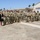 A large group of military personnel in camouflage uniforms are assembled outdoors on a paved area, likely at a military installation. One civilian woman in a black dress stands among the uniformed personnel, suggesting a formal ceremony or official event. The setting appears to be a tropical or subtropical location based on the palm trees and building architecture visible in the background.
