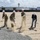 A groundbreaking ceremony is taking place at what appears to be a military installation, with six participants simultaneously shoveling dirt. The group includes both military personnel in camouflage uniforms and civilians in business attire, all wearing white hard hats. An air traffic control tower and low military-style buildings are visible in the background.