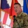 A U.S. Air Force military officer in OCP (Operational Camouflage Pattern) uniform is speaking at a podium with a microphone during what appears to be an official ceremony. Multiple flags are visible in the background, including the U.S. flag, what appears to be a state flag, and a blue Air Force flag. The event is being held under a tent or canopy structure.