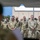 A group of military personnel in OCP (Operational Camouflage Pattern) uniforms are standing in formation, appearing to attend a formal ceremony or briefing outdoors. Several individuals are wearing face masks, suggesting this was taken during the COVID-19 pandemic period. The setting appears to be a military installation with a hangar or building visible in the background.