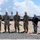 A military groundbreaking ceremony featuring six individuals — four in military uniforms and two in civilian business attire — all wearing white hard hats and holding ceremonial gold shovels. The event appears to take place on a sandy construction site with a control tower and military/airfield infrastructure visible in the background. The setting and uniforms suggest a U.S. military installation construction project.