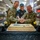 247th Army birthday cake cutting ceremony Three individuals — two in U.S. Army combat uniforms and one in civilian attire wearing a cap with a rank insignia — are jointly cutting a large sheet cake decorated with 'Army Birthday' text and gold frosting. The event appears to be an Army Birthday celebration held in a military dining facility (DFAC). Black and gold balloons and decorative elements reinforce the celebratory theme.
